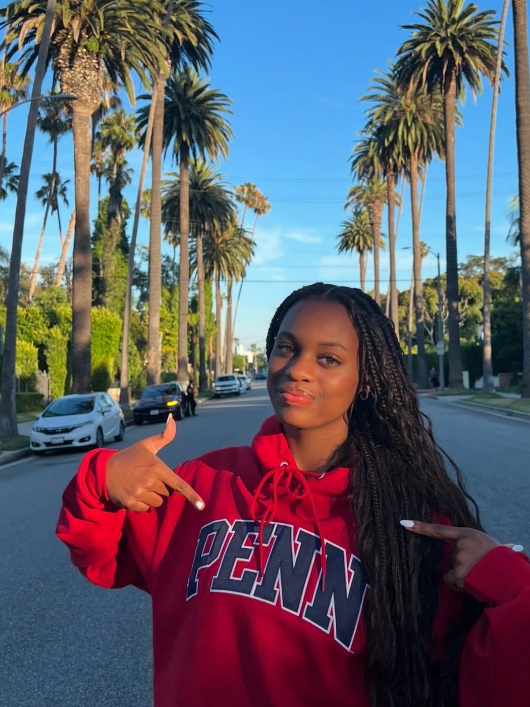 Lauryn Lewis in a red Penn hoodie on a palm lined Los Angeles street, pointing at the Penn lettering.